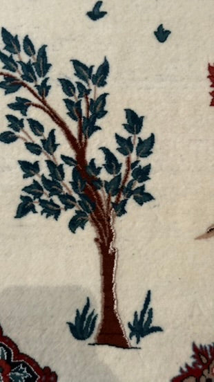A close-up of a cream-colored rug featuring a stylized tree motif with dark blue leaves and a brown trunk, surrounded by small birds and floral details.