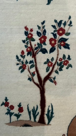 A close-up of a woven textile featuring a stylized tree motif with red flowers, dark green leaves, and a brown trunk on an off-white background.