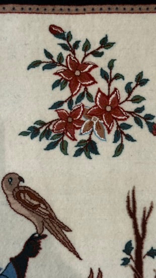Close-up of a woven textile or rug pattern featuring red flowers with green leaves on an off-white background, plus a brown bird and dark branch details.