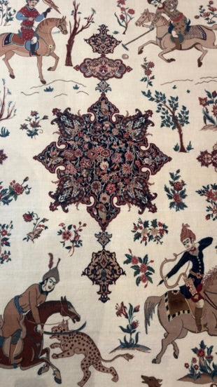 A close-up of a patterned textile or rug featuring a central ornate medallion surrounded by floral motifs and figures on horseback, with an off-white background and muted red, blue, brown, and black details.