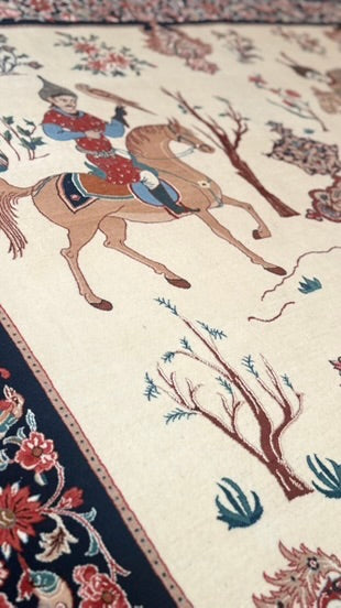 Close-up of an ornate rug with a cream background, featuring a central scene of a rider on a horse in red and blue, surrounded by floral and tree motifs, with a dark navy border.