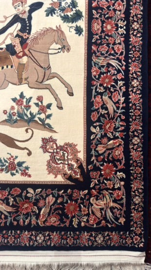 A close-up of an ornate patterned rug showing a cream field with a rider on horseback motif, surrounded by a dark navy floral and bird border with fringe at the bottom.