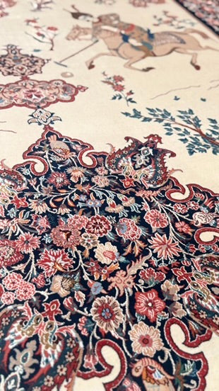 A close-up of an ornate patterned rug with a cream background and detailed floral, paisley, and horse motifs in red, navy, pink, and beige.