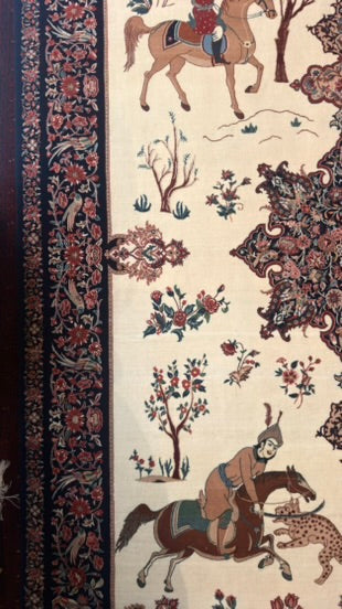 A close-up of an ornate patterned rug or textile with a cream background, dark navy border, and illustrated scenes of riders on horseback, trees, and floral motifs.