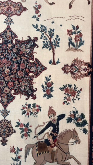 A close-up of an ornate rug or textile with a cream background, featuring floral motifs, trees, and a rider on horseback in dark blue, red, and brown tones.
