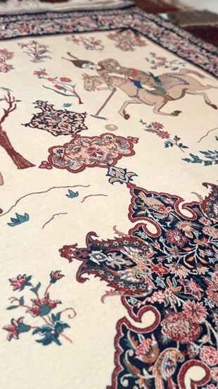 A close-up of an ornate patterned rug with a cream background, featuring traditional motifs including a horse rider, floral elements, and a dark navy border with red and pink detailing.