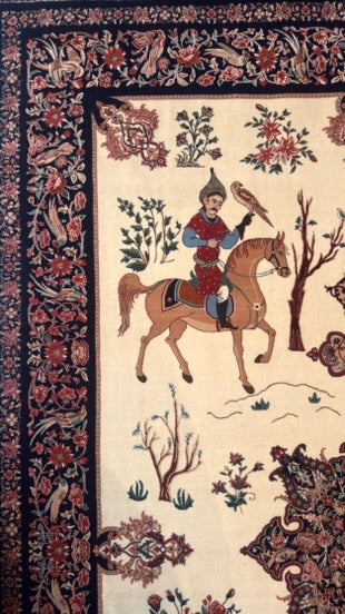 A close-up of an ornate rug featuring a central scene of a rider on a horse with a bird, surrounded by floral motifs and a dark patterned border.