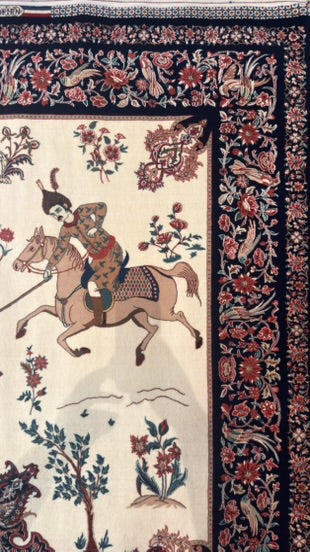 A close-up of an ornate patterned rug featuring a central scene of a rider on horseback, surrounded by a dark floral border on a cream background.