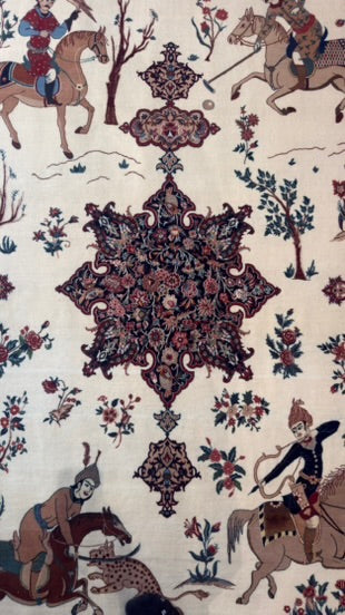 A close-up of an ornate patterned textile or rug featuring a central medallion and surrounding motifs of riders on horseback, floral elements, and trees in muted red, blue, green, and brown on a light beige background.