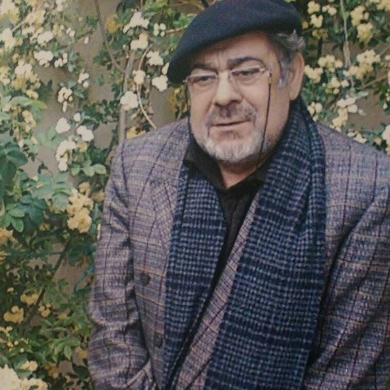 Man wearing a checkered jacket and beret standing in front of floral bushes